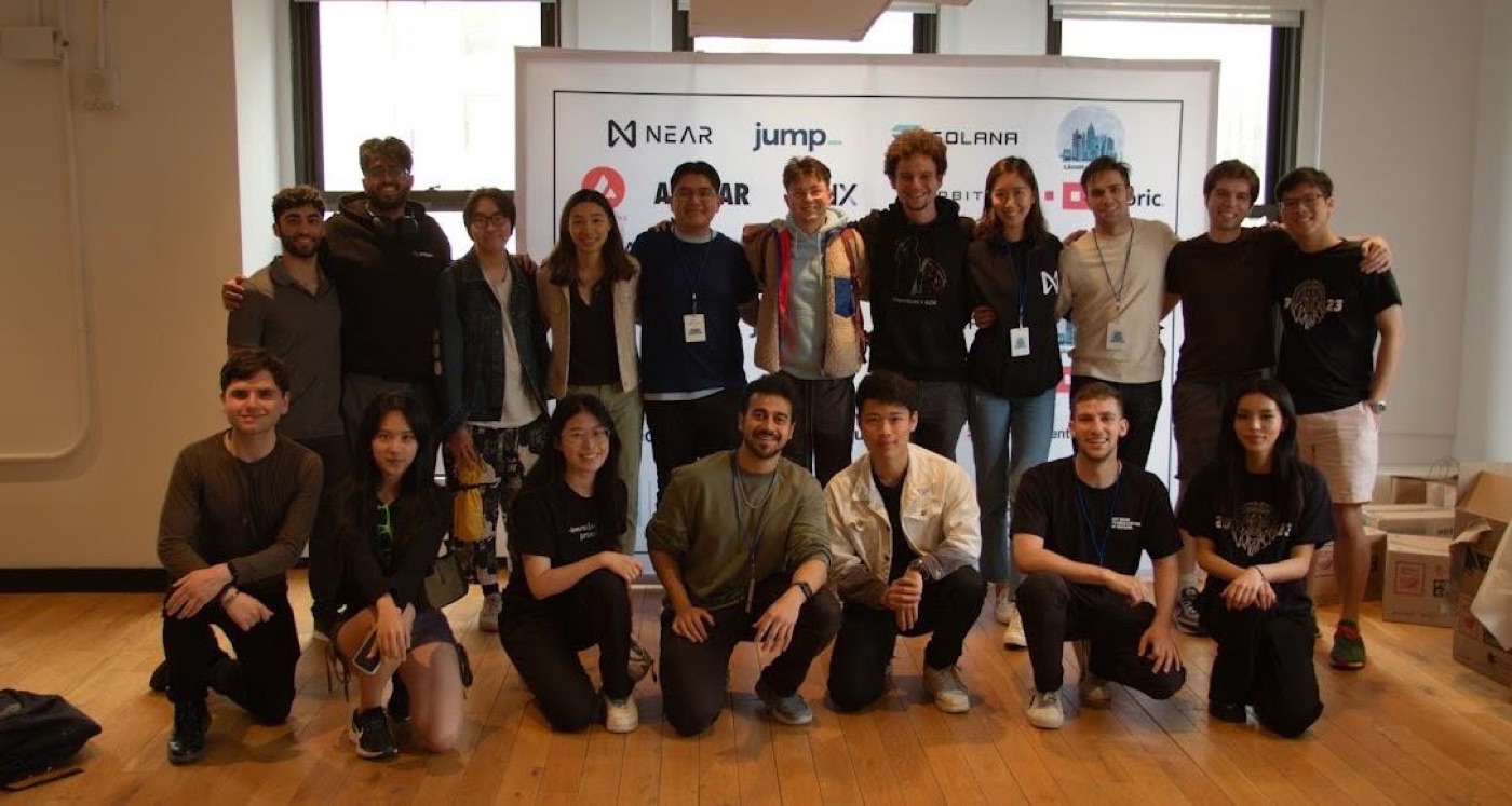 About twenty hackathon participants pose in two rows in front of a sponsor banner in a sunlit, wood-floored room.