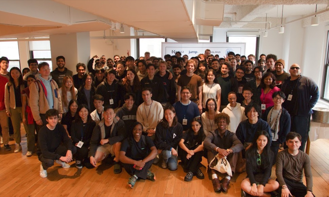 A crowd of hackathon attendees, close to a hundred, packs a bright loft room in front of the sponsor backdrop.