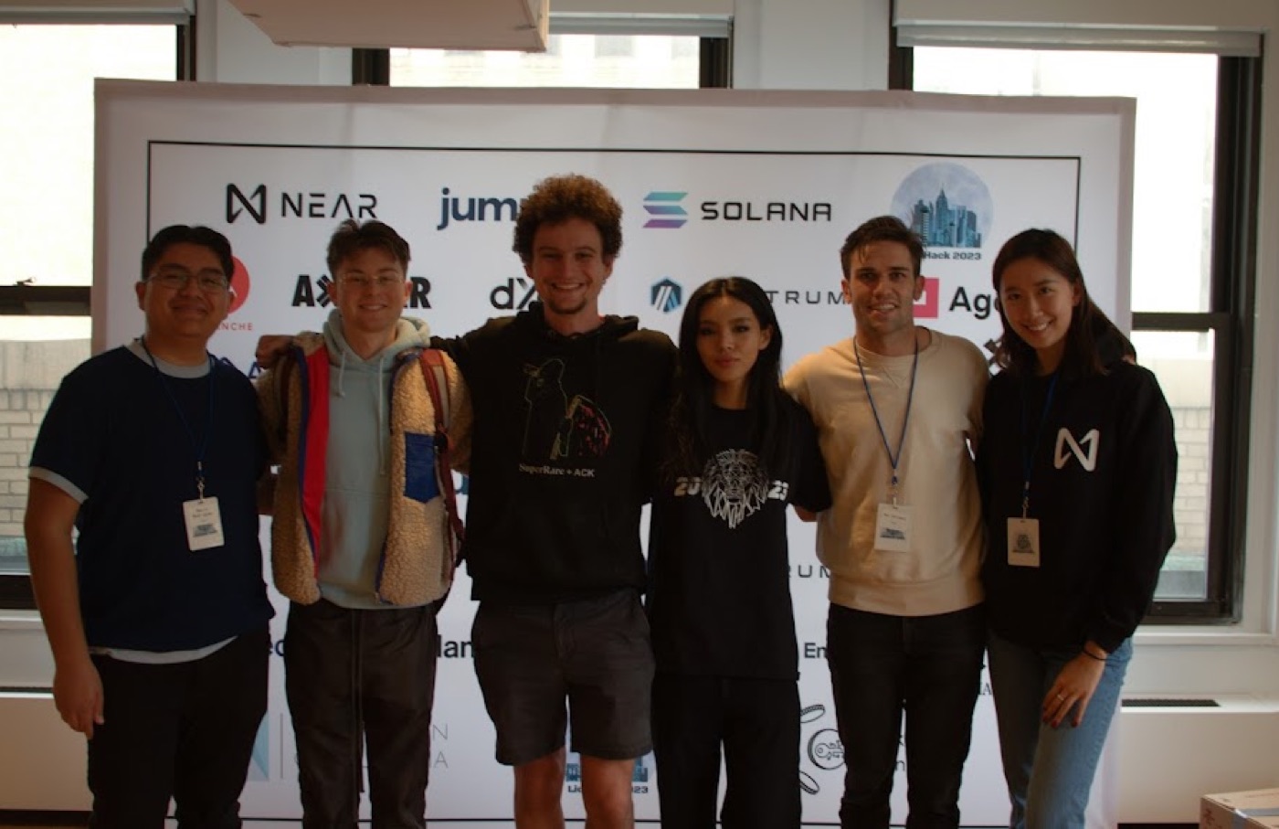Six hackathon attendees with lanyards line up arm-over-shoulder in front of the sponsor wall between two windows.