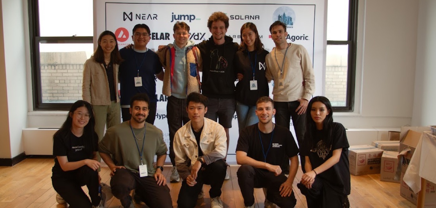 Eleven hackathon attendees pose in two rows before the sponsor banner, packing boxes stacked at the edges of the room.