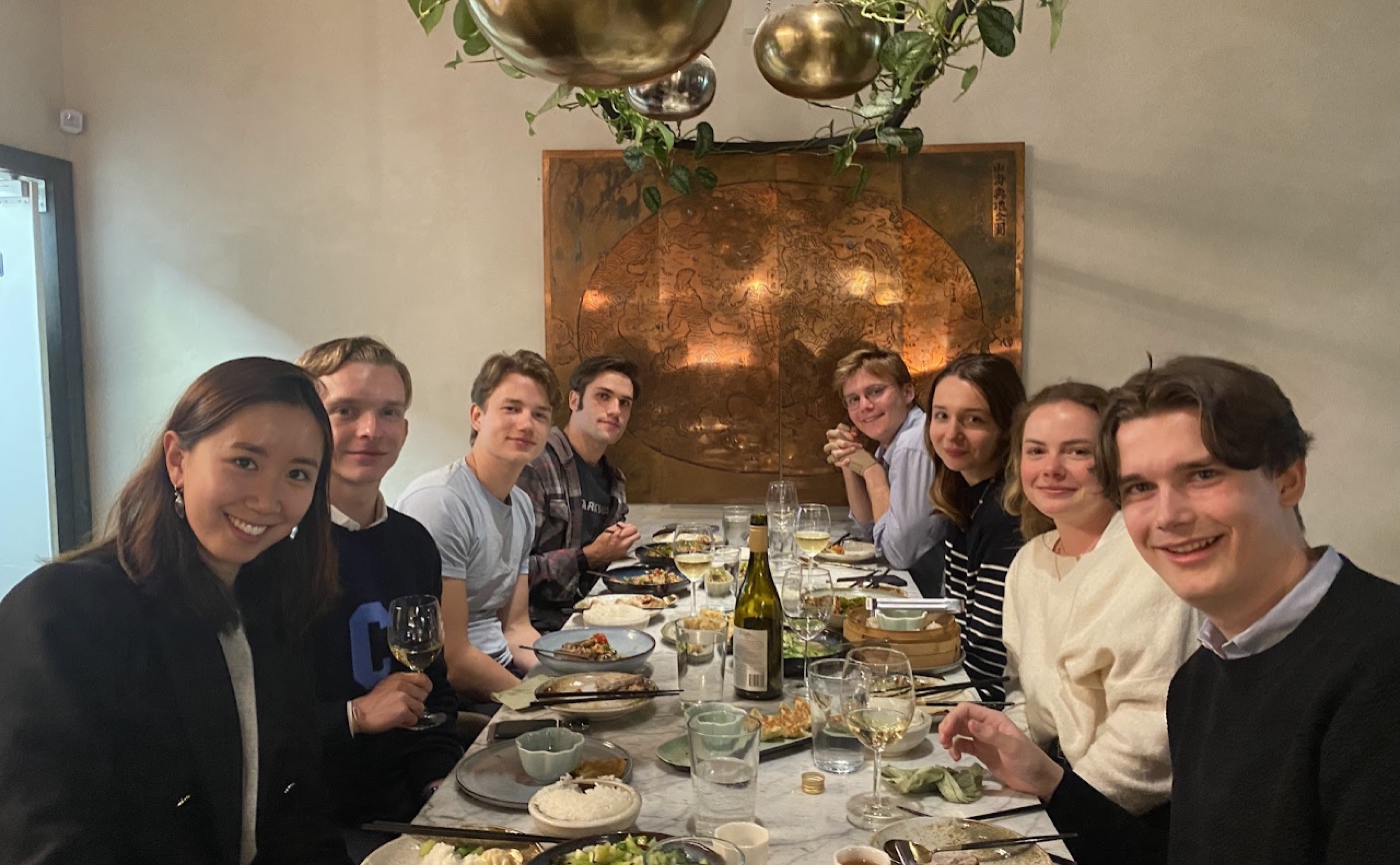Eight friends at a marble dinner table look up from dumplings and rice, brass pendant globes hanging overhead.