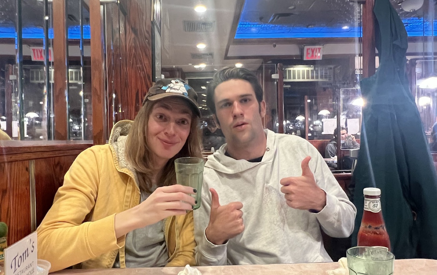 Noel gives two thumbs up in a white hoodie beside a long-haired friend raising a green diner glass at Tom's Restaurant.
