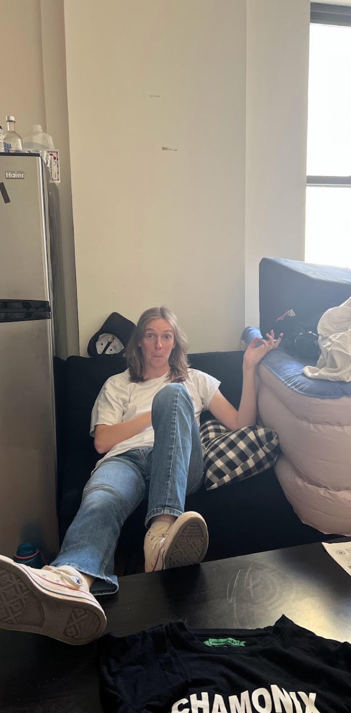 A long-haired friend sprawls on a black couch beside a mini-fridge, mid-shrug, in a sunlit apartment.