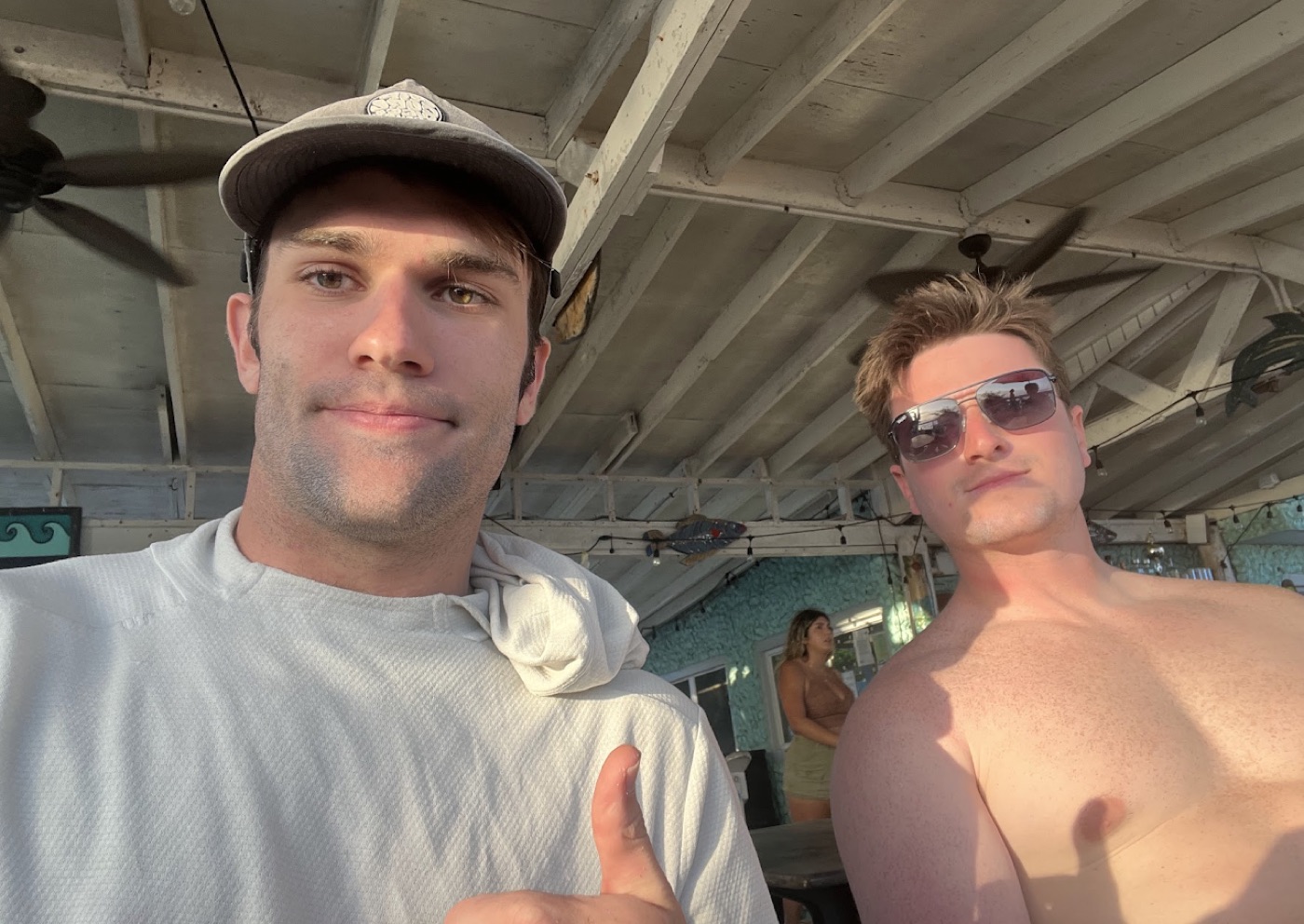 Selfie of Noel in a gray cap giving a thumbs-up beside a sunburned friend in aviators under the white rafters of an open-air beach bar.