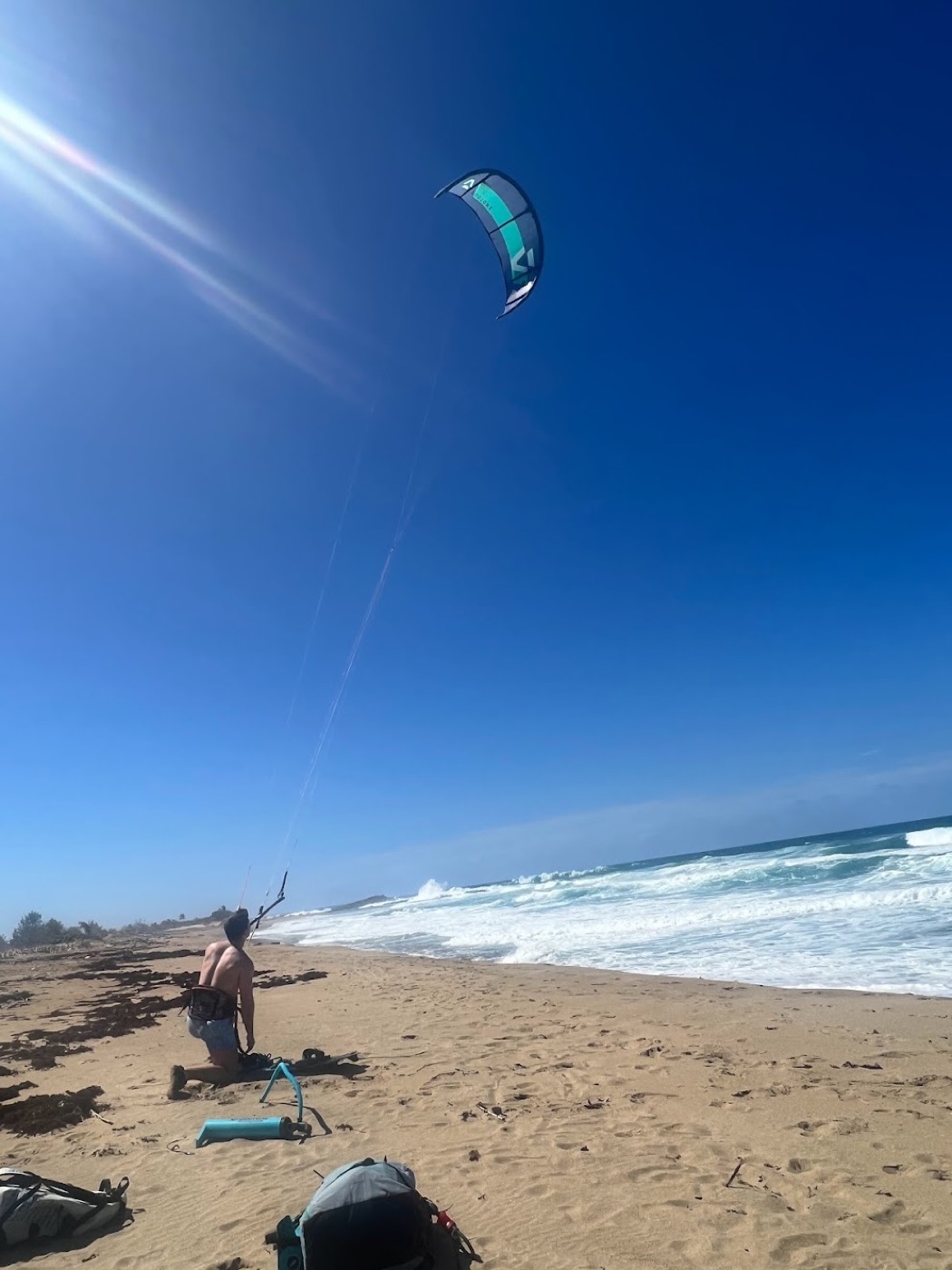 A kiteboarder kneels on an empty beach steering a blue-and-teal kite high over whitecapped surf under a deep blue sky.