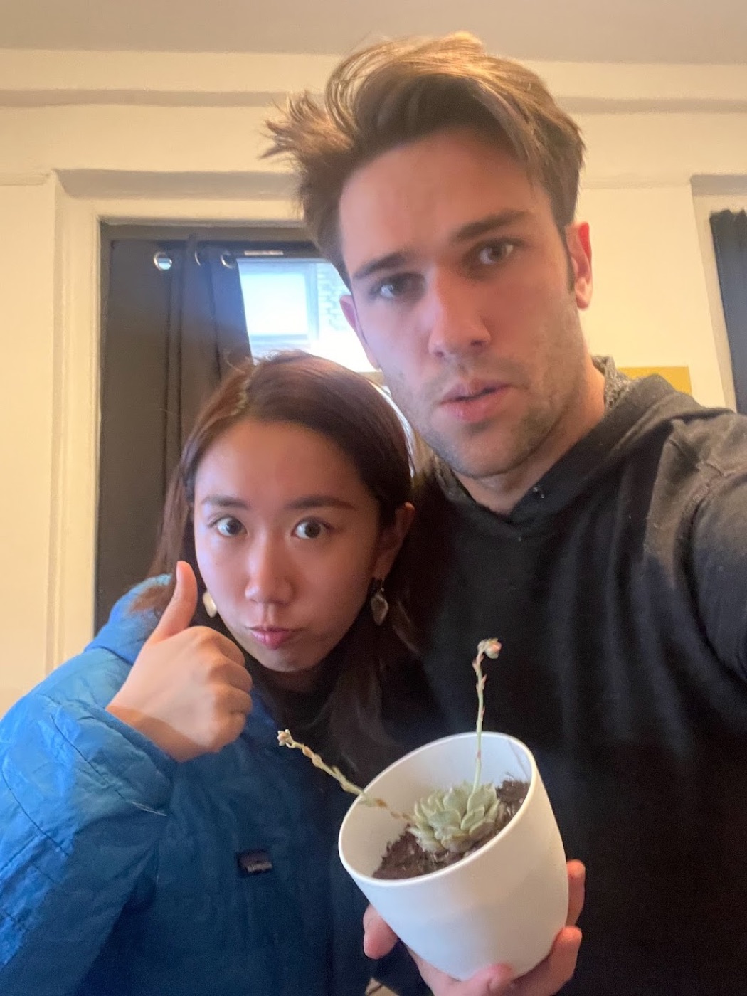 Selfie of Noel and a woman giving deadpan thumbs-ups over a struggling potted succulent with two leggy stems.