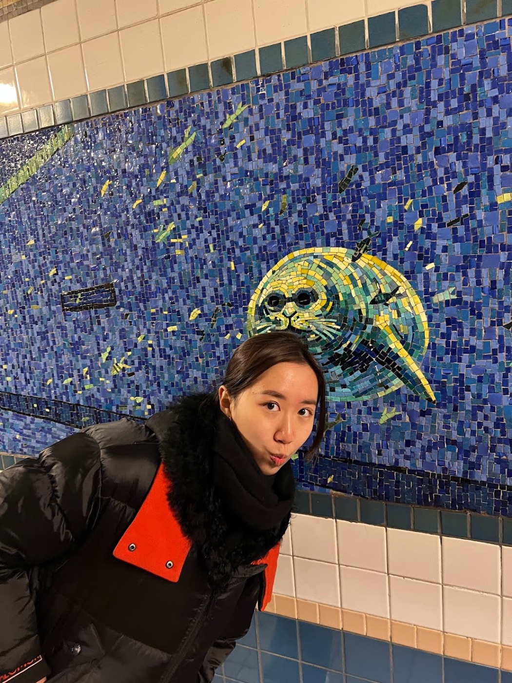 A woman puckers at the camera beneath a blue subway mosaic of a wide-eyed seal in a tiled station corridor.