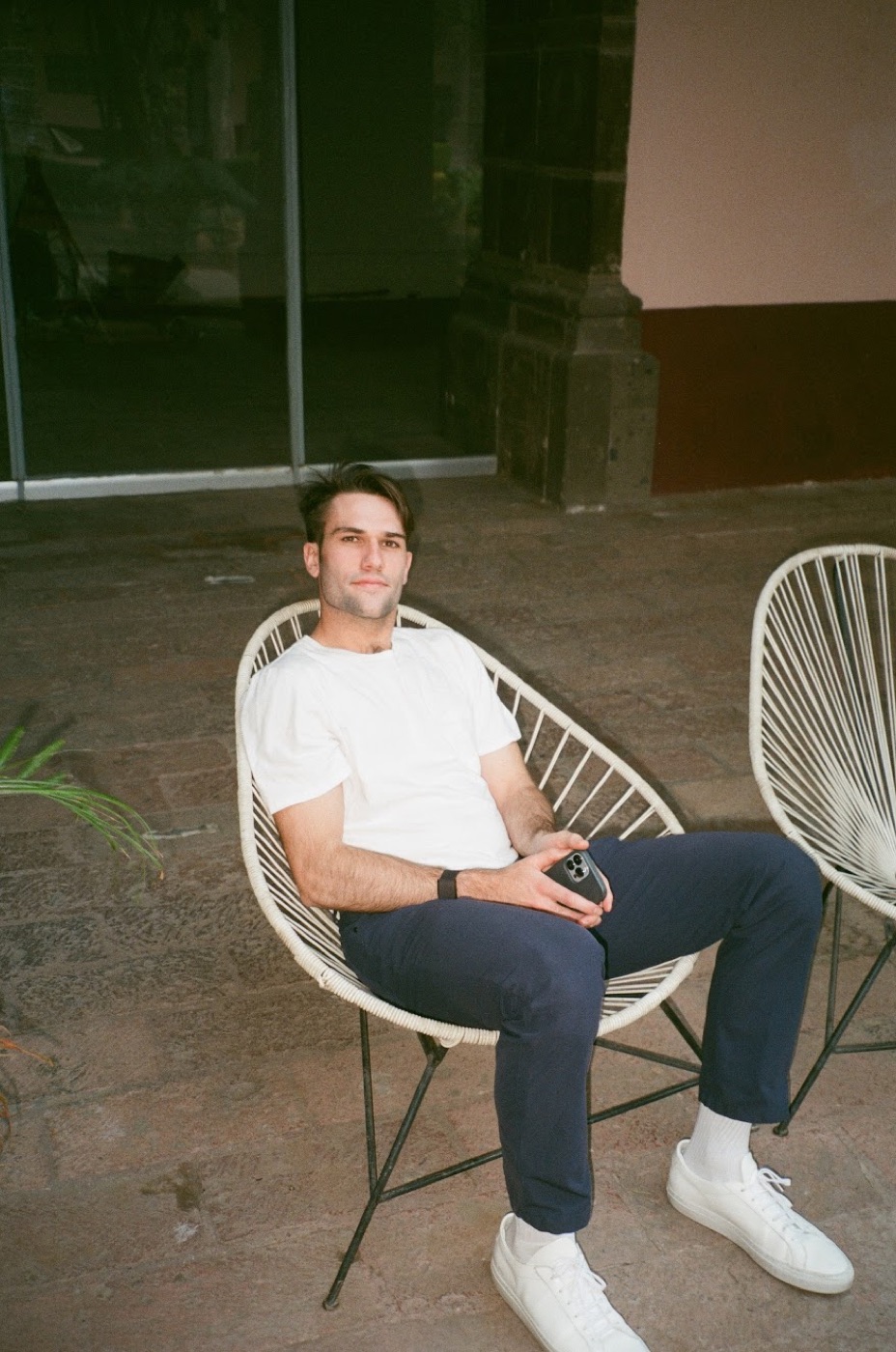 Film photo of Noel reclining in a white woven chair on a stone patio, phone in hand, looking up at the camera.