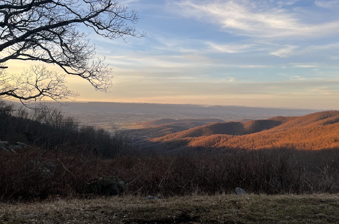 Golden-hour view from a mountain overlook, low sun lighting wooded ridgelines above a patchwork valley.