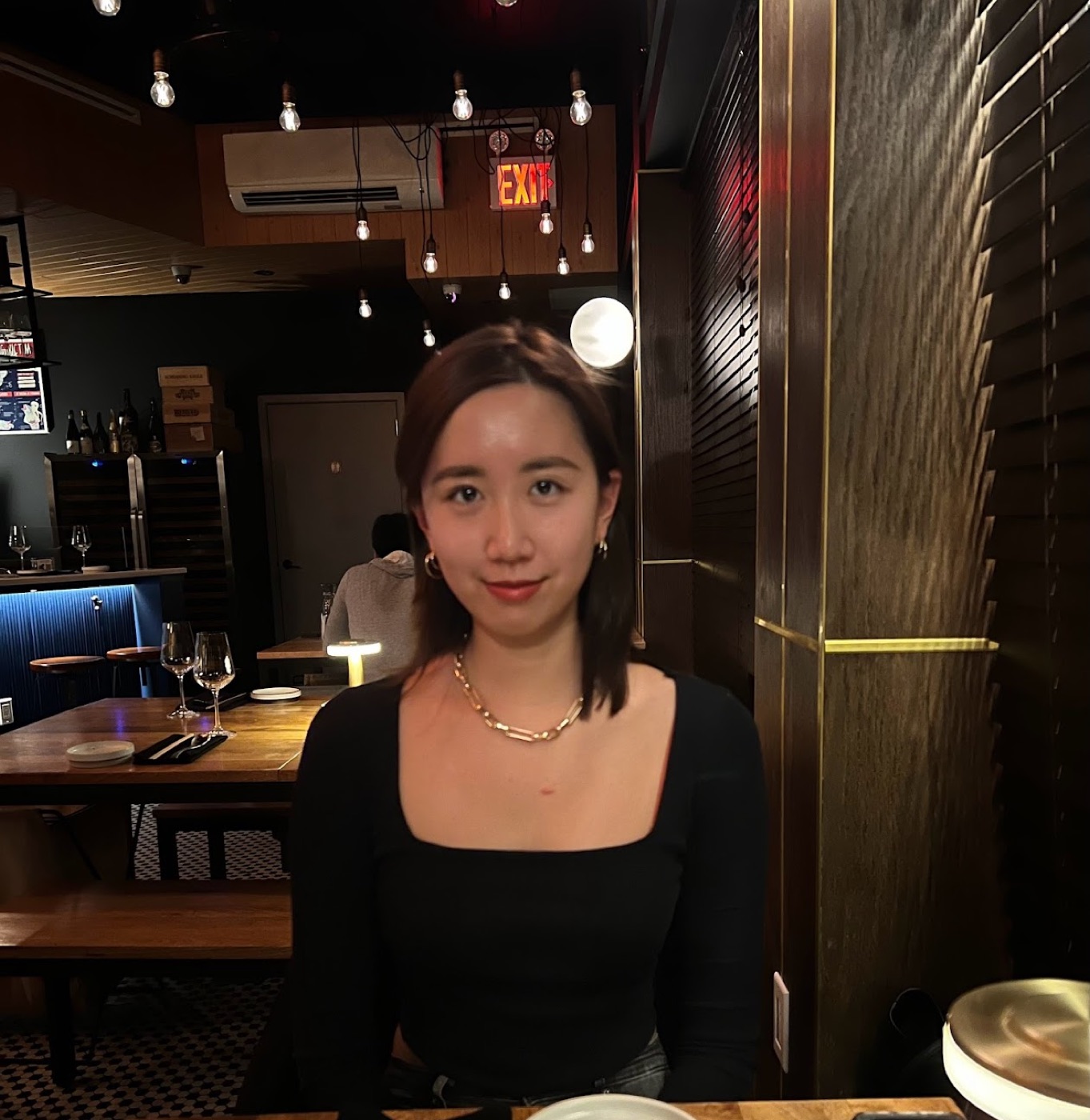 A woman in a black square-neck top smiles softly at a table in a dark bar strung with exposed bulbs, an EXIT sign glowing behind her.