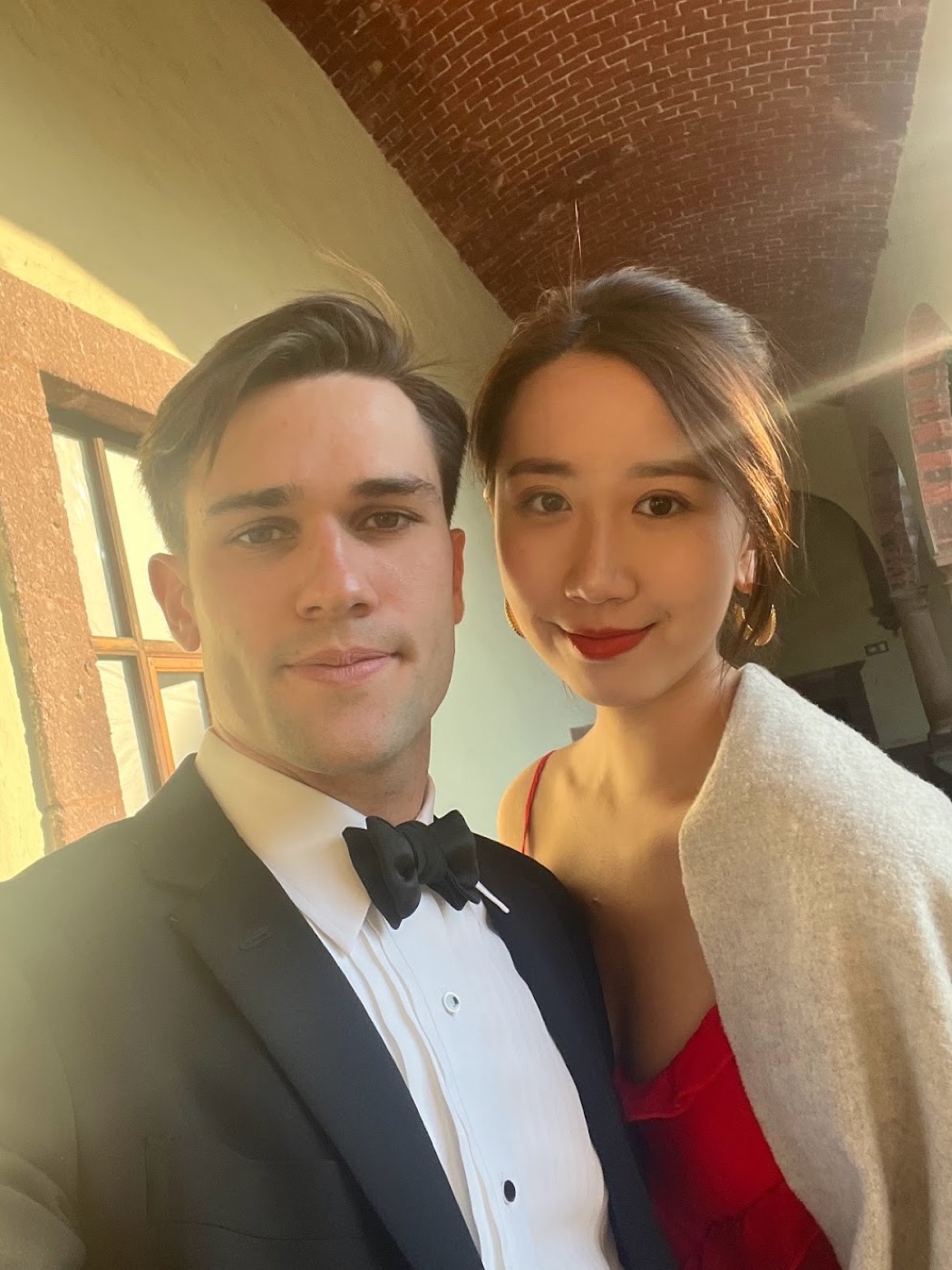Selfie of Noel in a tuxedo and a woman in a red dress with a cream shawl beneath a vaulted brick ceiling.