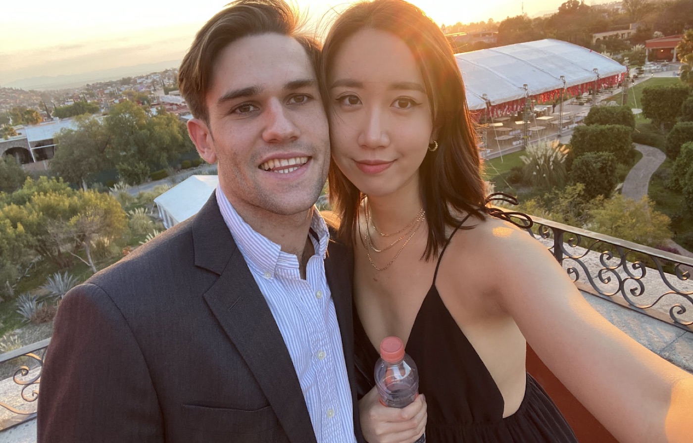 Sunset selfie of Noel in a suit and a woman in a black dress on a terrace above gardens and a large white event tent.