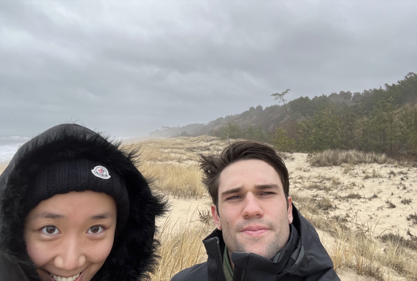 Windblown winter-beach selfie of a woman in a fur-lined hood and Noel in a parka, grassy dunes under a gray sky behind them.